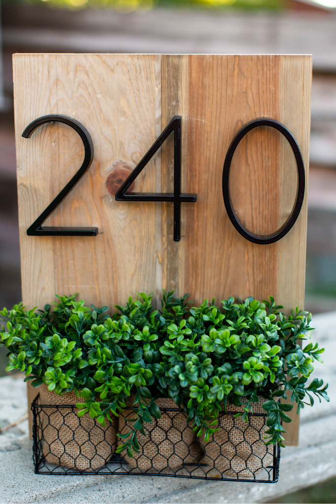 Black zinc alloy house numbers 2, 4 and 0 on a wood plaque above greenery